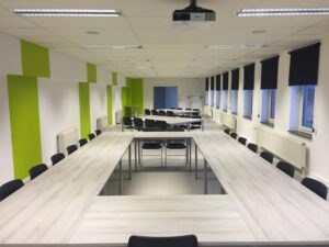 a large classroom with a hanging projector and chairs around long tables